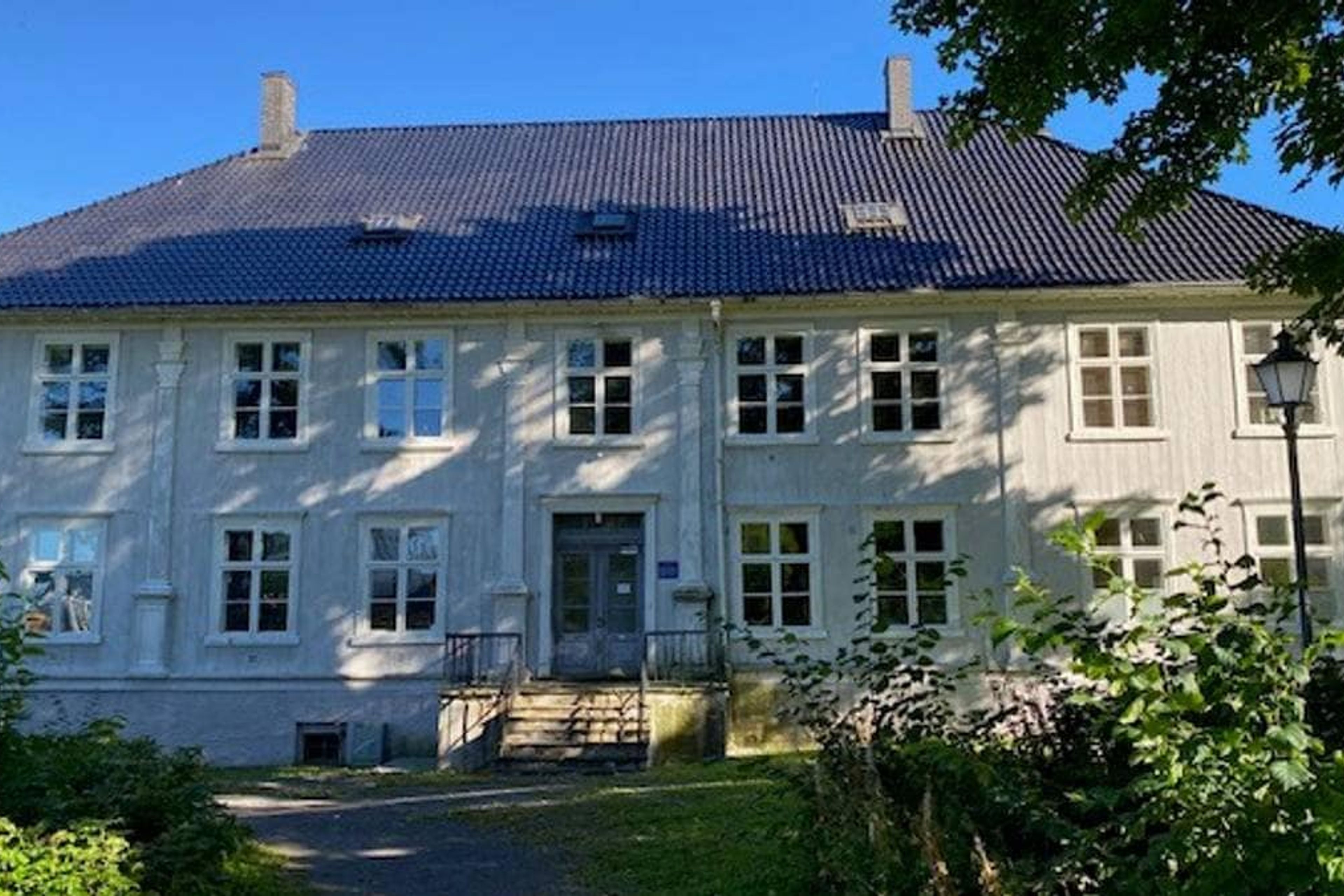En hvit, ærverdig murbygning med blått tak omgitt av grønt – mottaket i Holmestrand.