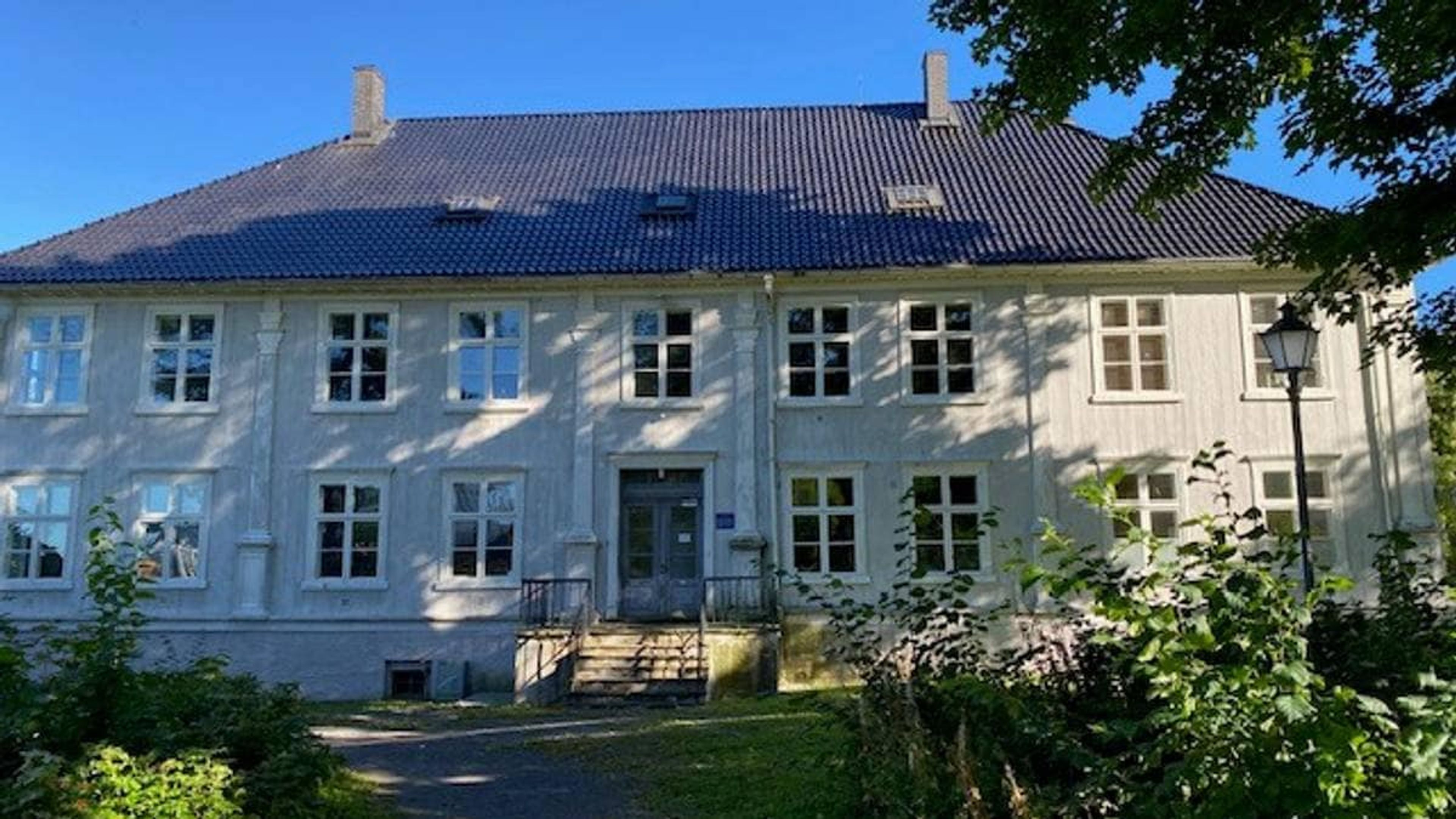 En hvit, ærverdig murbygning med blått tak omgitt av grønt – mottaket i Holmestrand.