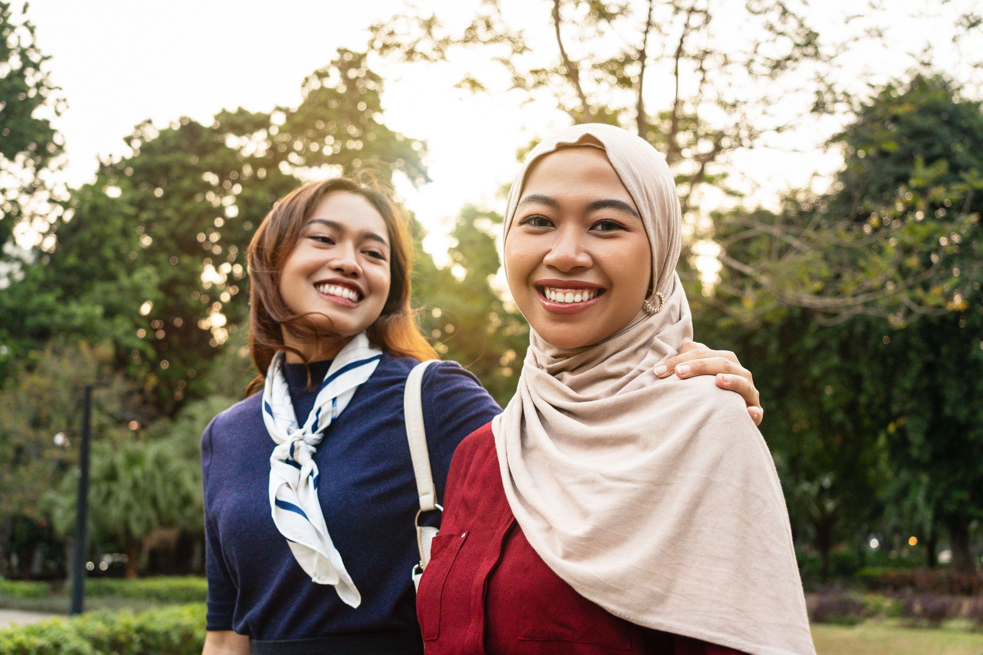 To smilende unge kvinner utendørs i en park, den ene med hijab.