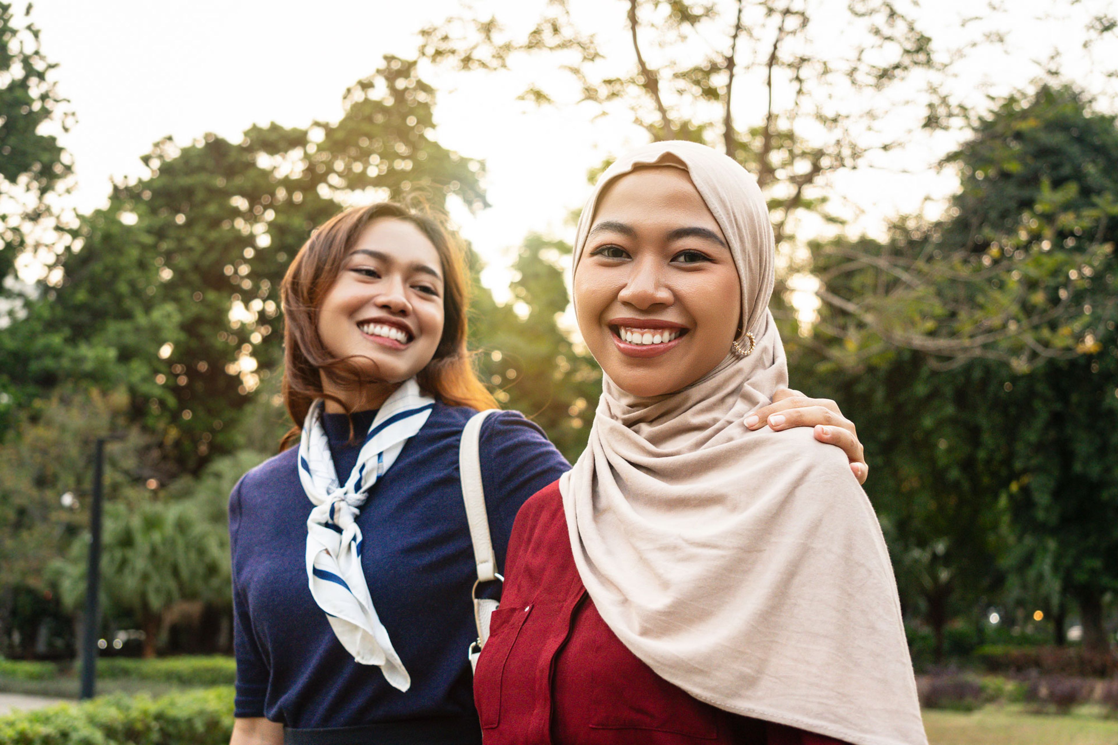 To smilende unge kvinner utendørs i en park, den ene med hijab.