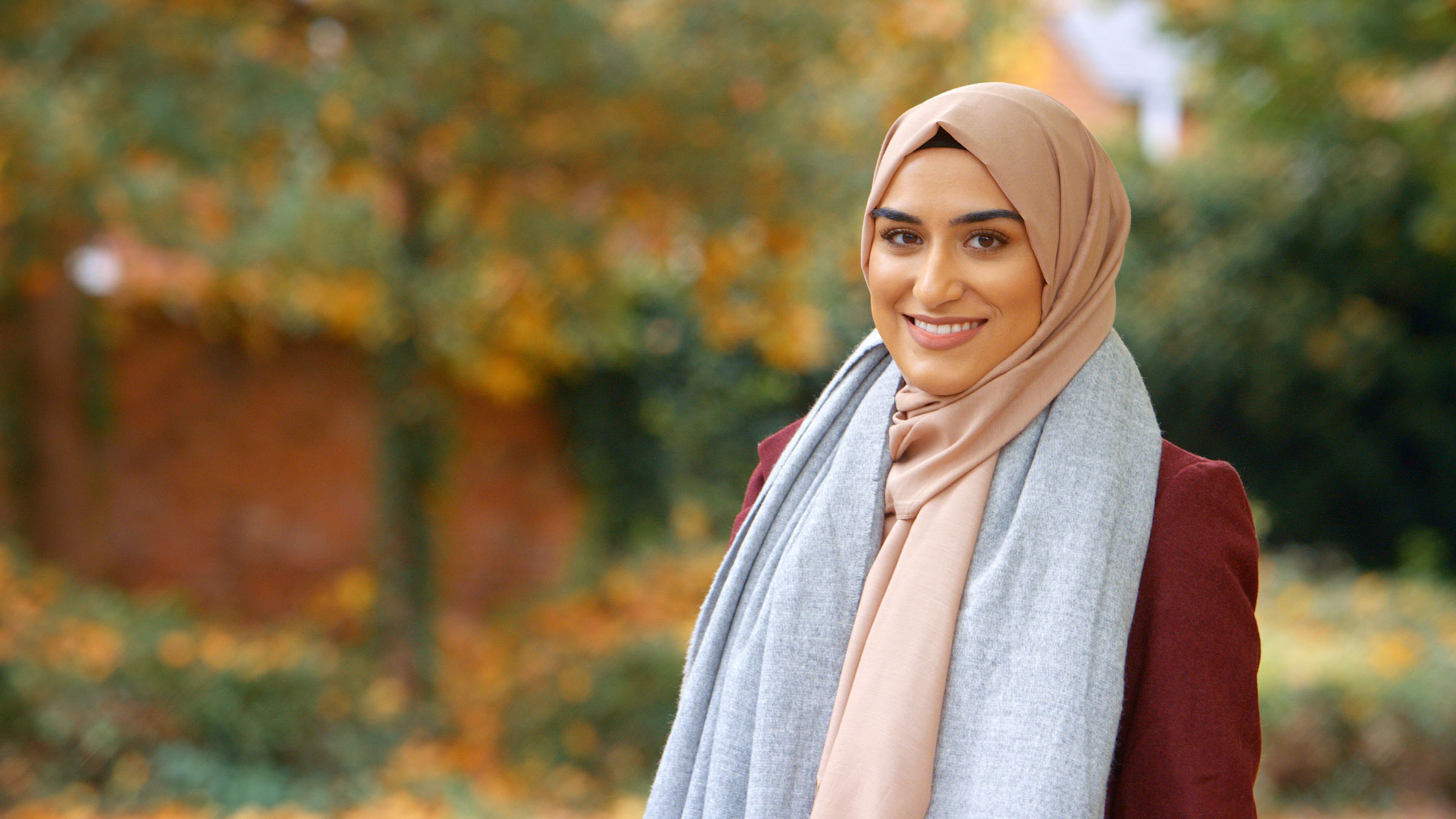 Portrett av en smilende kvinne med hijab utendørs i høstlige omgivelser.
