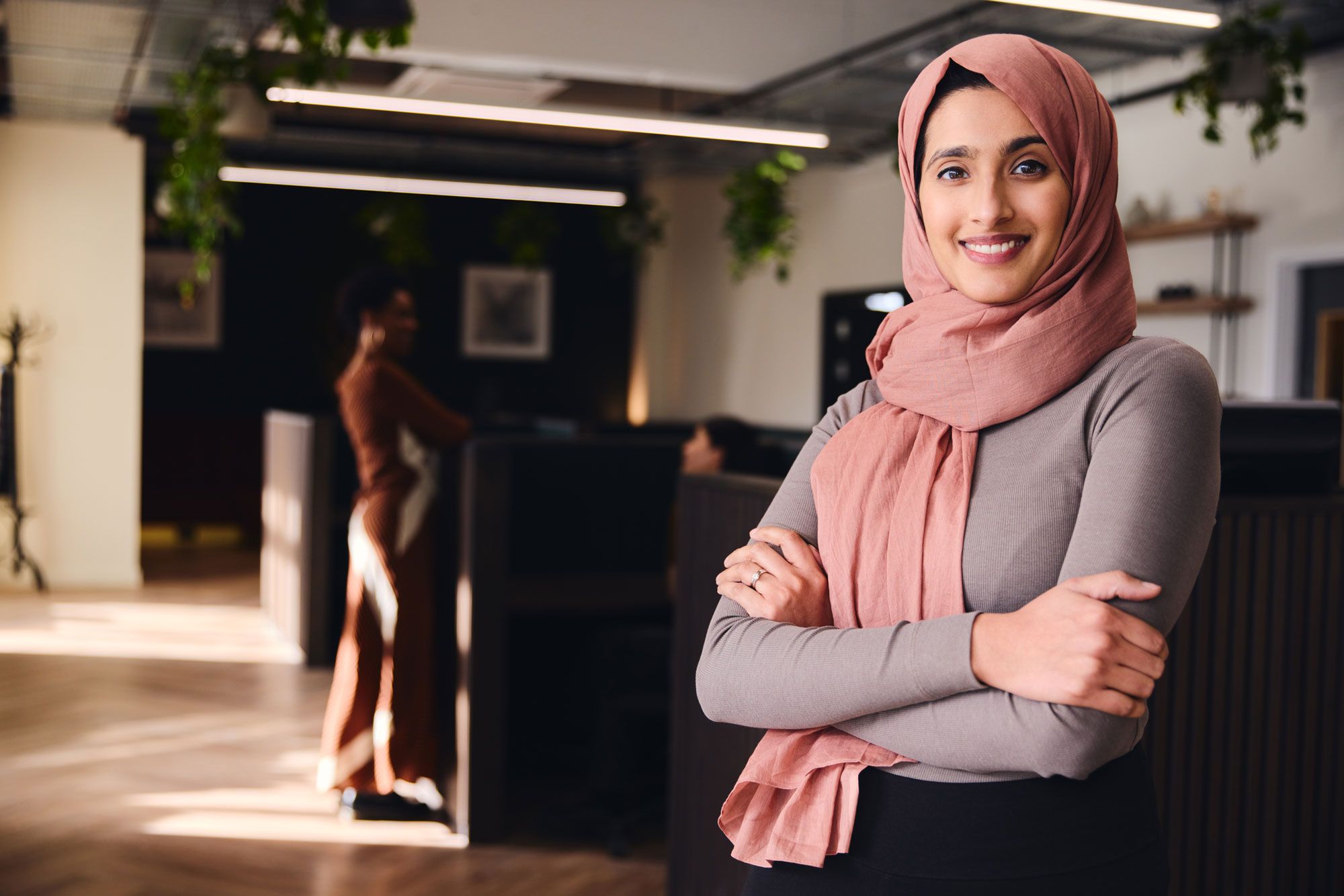 Smilende kvinne med rosa hijab står med armene i kors i et moderne kontorlokale.