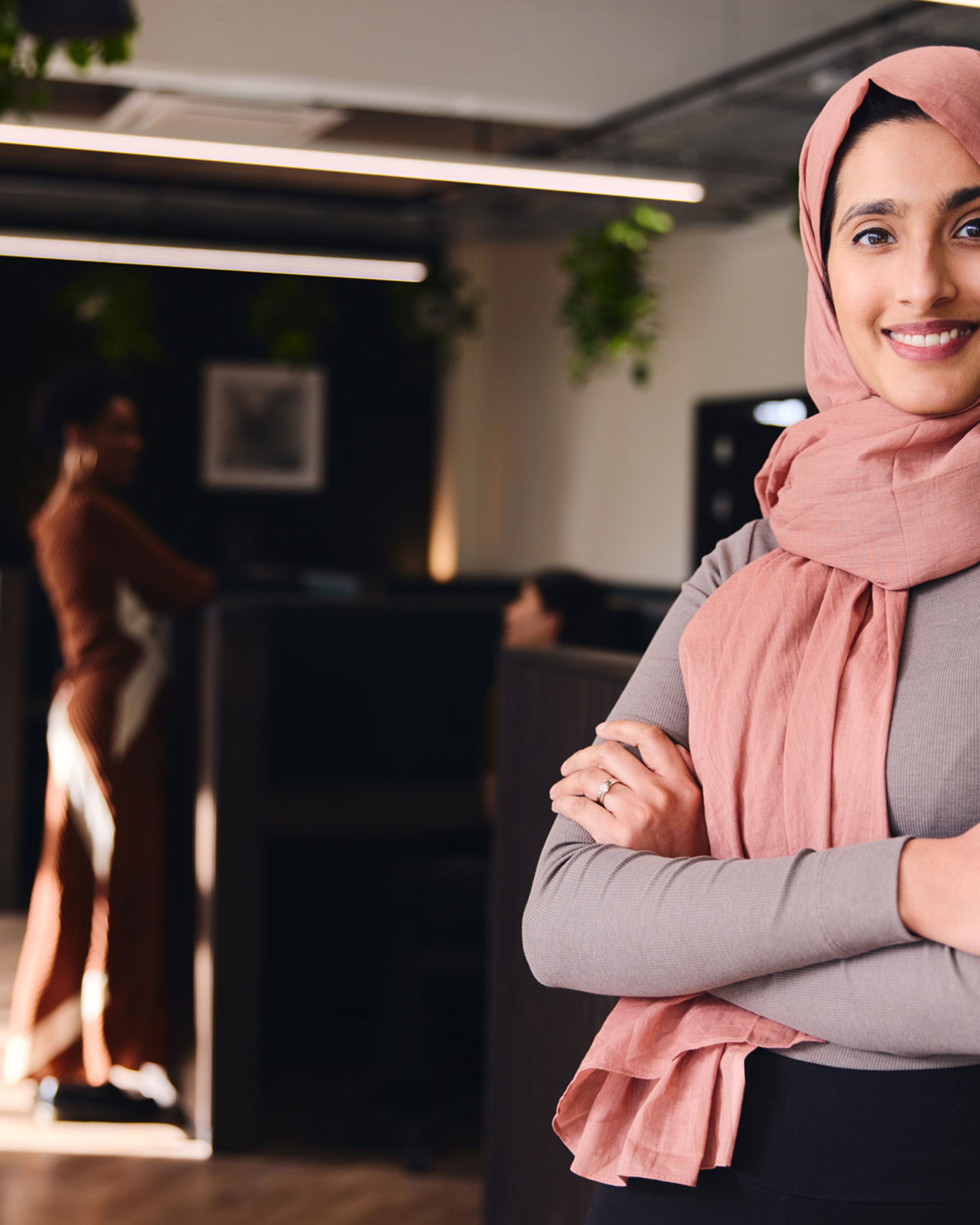 Smilende kvinne med rosa hijab står med armene i kors i et moderne kontorlokale.