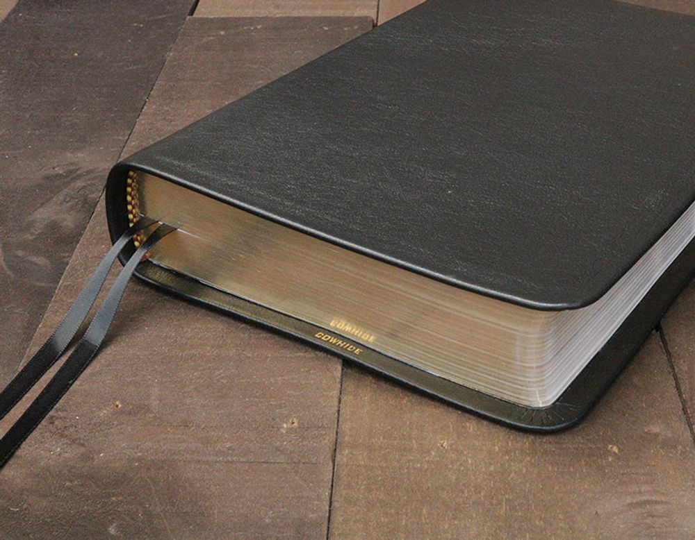 Black cowhide leather-bound Bible with gilded page edges and ribbon markers on a wood surface