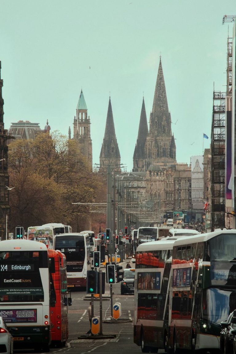 Busy city street in Edinburgh with red double-decker buses and tall church spires in the distance.