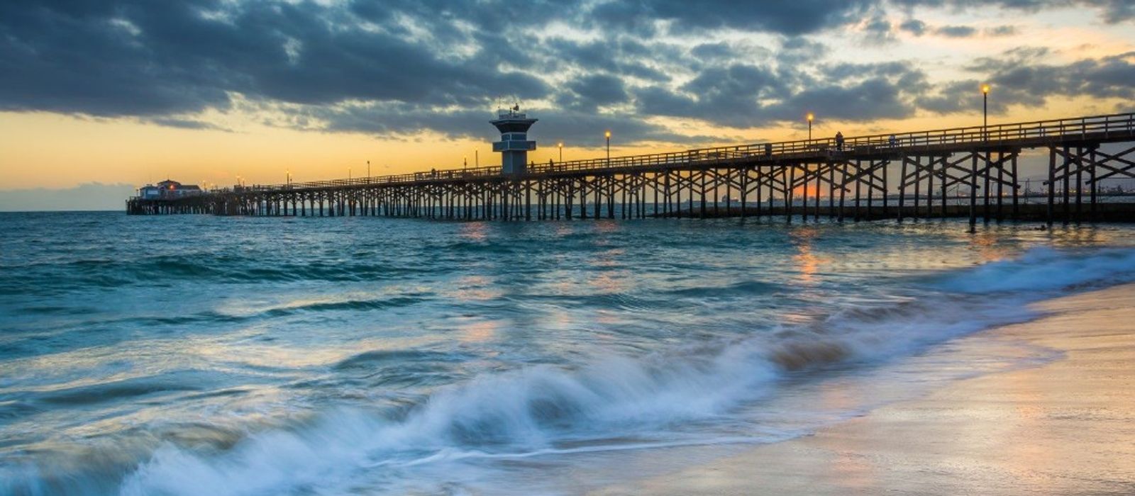 Seal Beach Pier at sunset with waves crashing on the shore and dramatic clouds over the Pacific Ocean