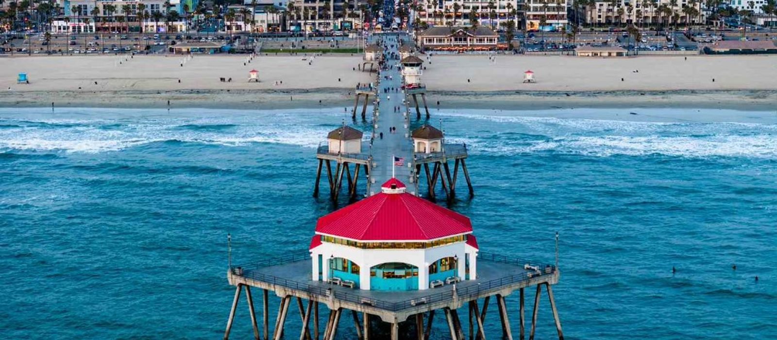 Aerial view of Huntington Beach Pier with its iconic red-roofed Ruby's Diner, the wide sandy beach, and Surf City skyline in the background