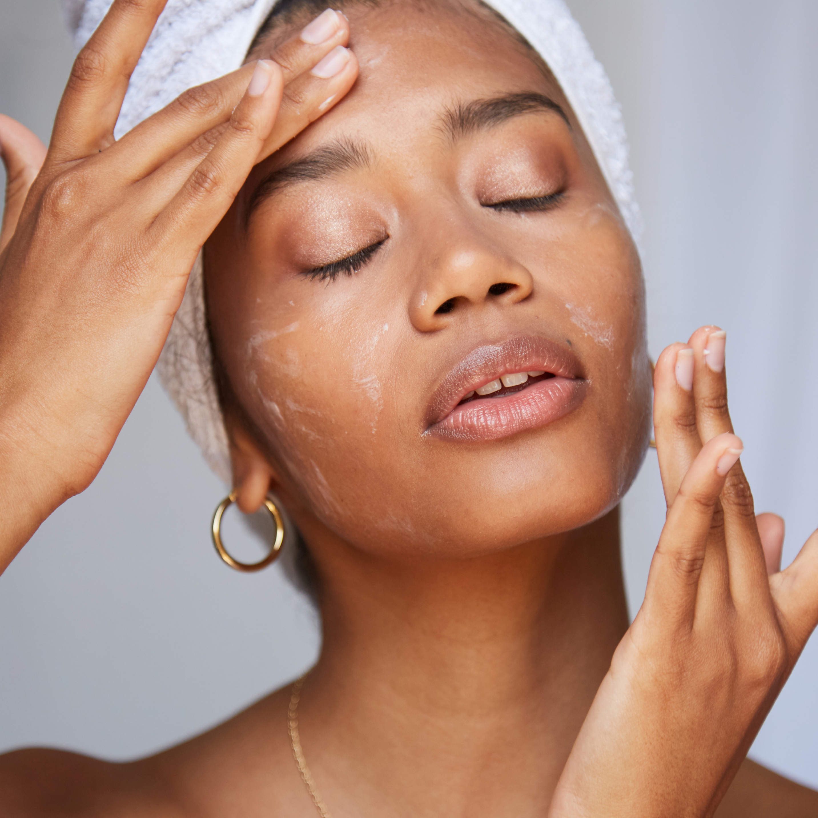 A model with her eyes closed and a towel on her hair applying Physiogel Daily Moisture Therapy Intensive Cream to her face