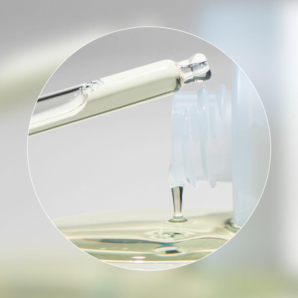 Close-up image of a glass dropper dispersing oil from a clear glass bottle. The dropper is releasing a drop of clearliquid into the bottle, with a smooth and shiny texture evident. The background is blurred, focusing attention on the dropper and bottle.