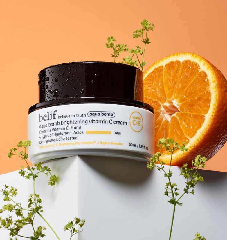 White jar of belif Aqua Bomb Brightening Vitamin C Cream standing on a white block with half of orange fruit and green sprigs by its side.