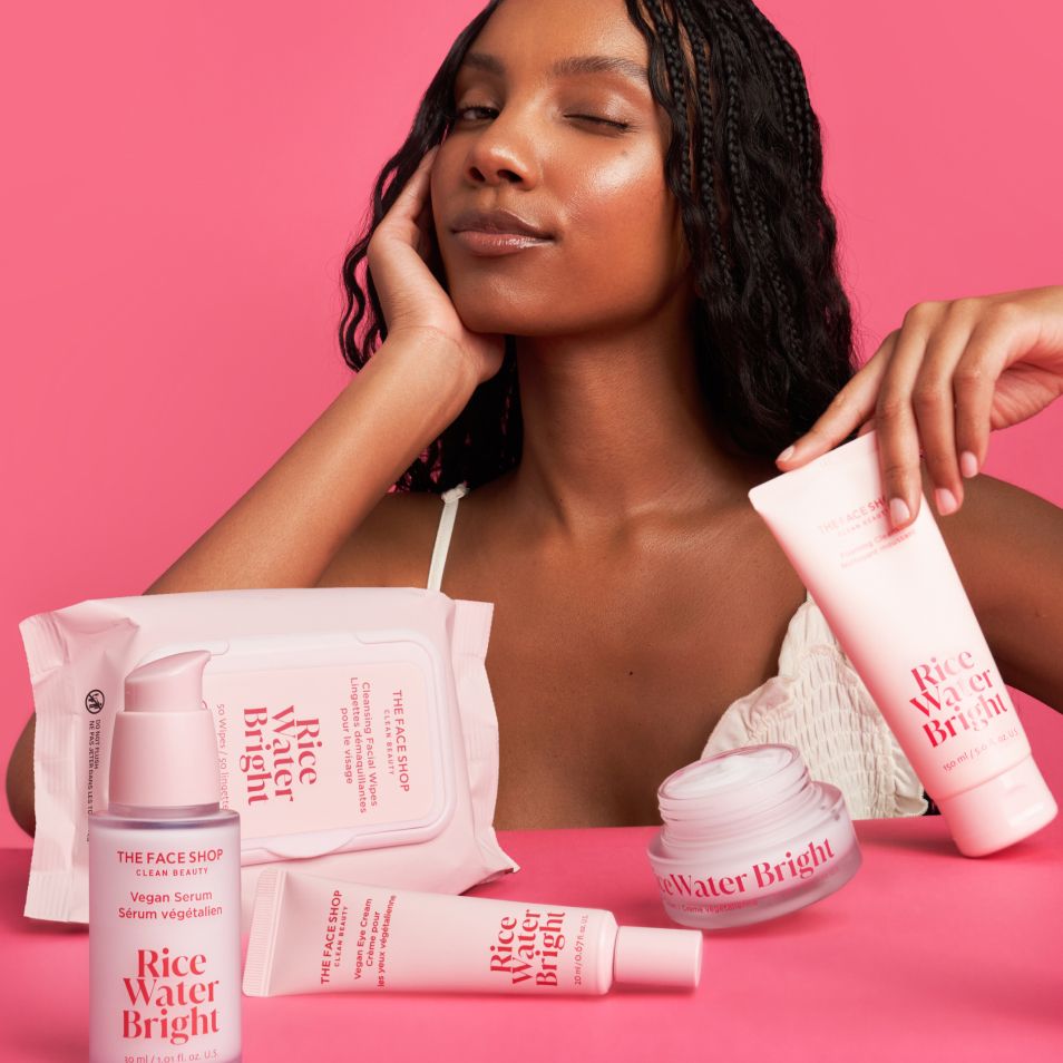 A winking woman with glowing skin poses with pink Rice Water Bright skincare products on a pink background.