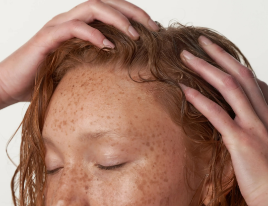 A woman with red hair has her eyes closed and has her hands holding her freshly washed hair on top of her head.