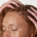 A woman with red hair has her eyes closed and has her hands holding her freshly washed hair on top of her head.