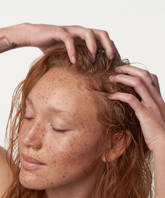 Close-up of a person with freckled skin and wet, reddish-brown hair, eyes closed, gently massaging their scalp.