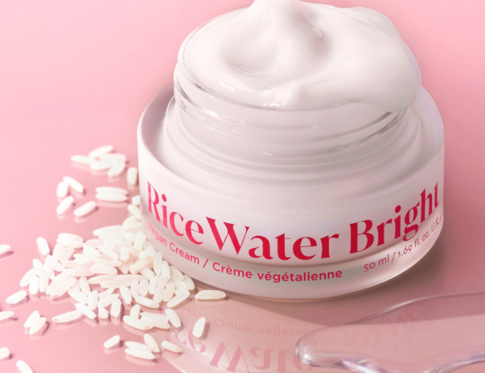 A jar of The Face Shop's Rice Water Bright Vegan Cream overflowing with white product, surrounded by rice grains, on a pink background.