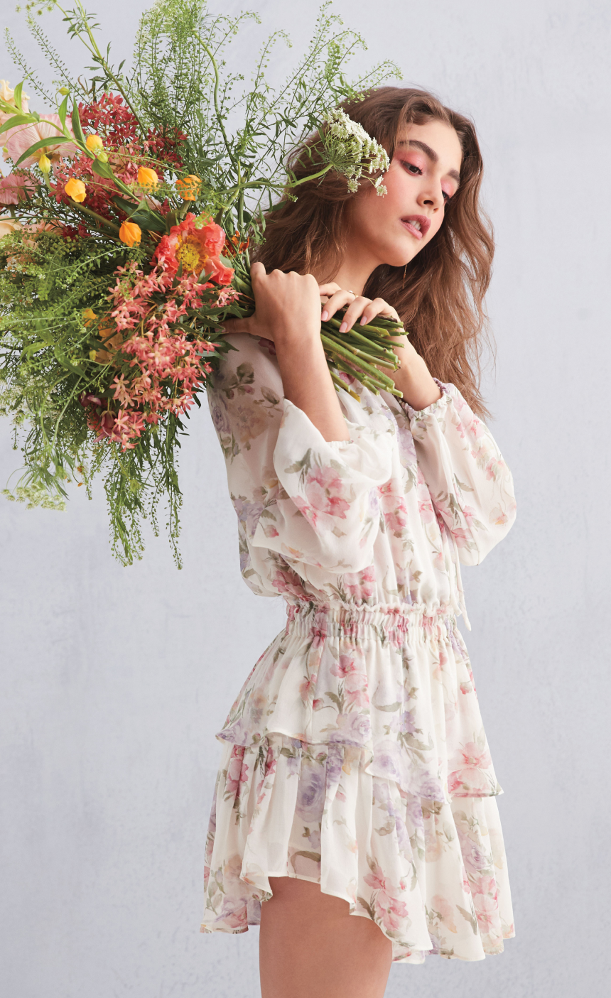 A model in a floral print dress holding a bouquet of flowers to her shoulder.