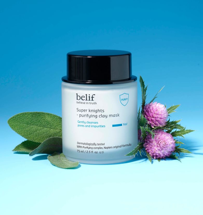 A white jar of belif Super Knights - Purifying Clay Mask standing on a light blue surface with a pink herb lying around.