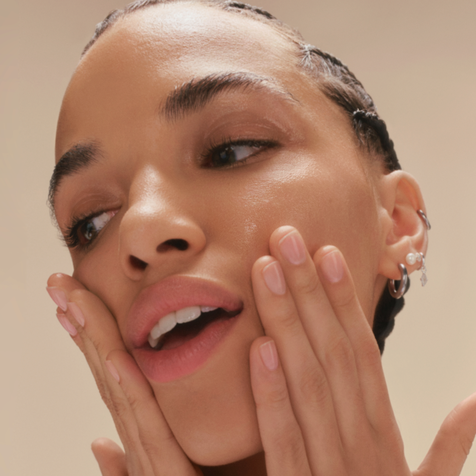 Close-up portrait of a smiling young woman with braids with both hands on her face rubbing in a skincare product on a warm beige background.