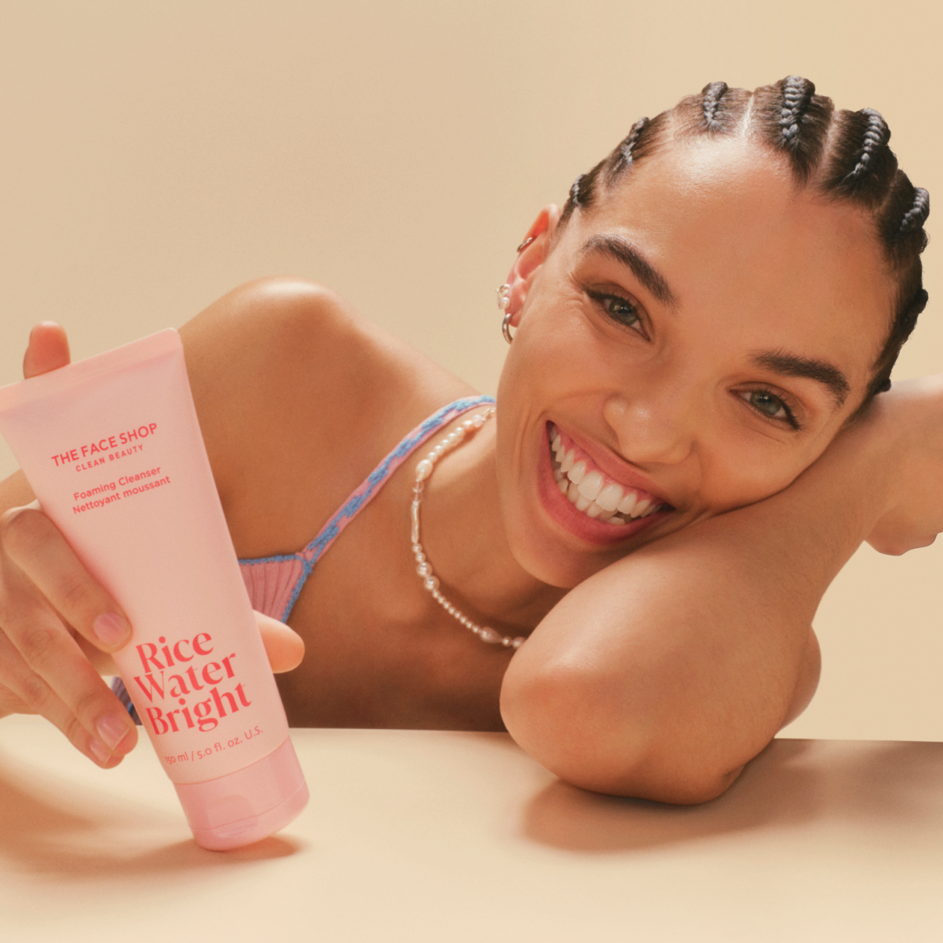 A joyful woman with cornrow braids smiles warmly, holding up a pink bottle of the face shop rice water bright foaming cleanser against a soft beige background.