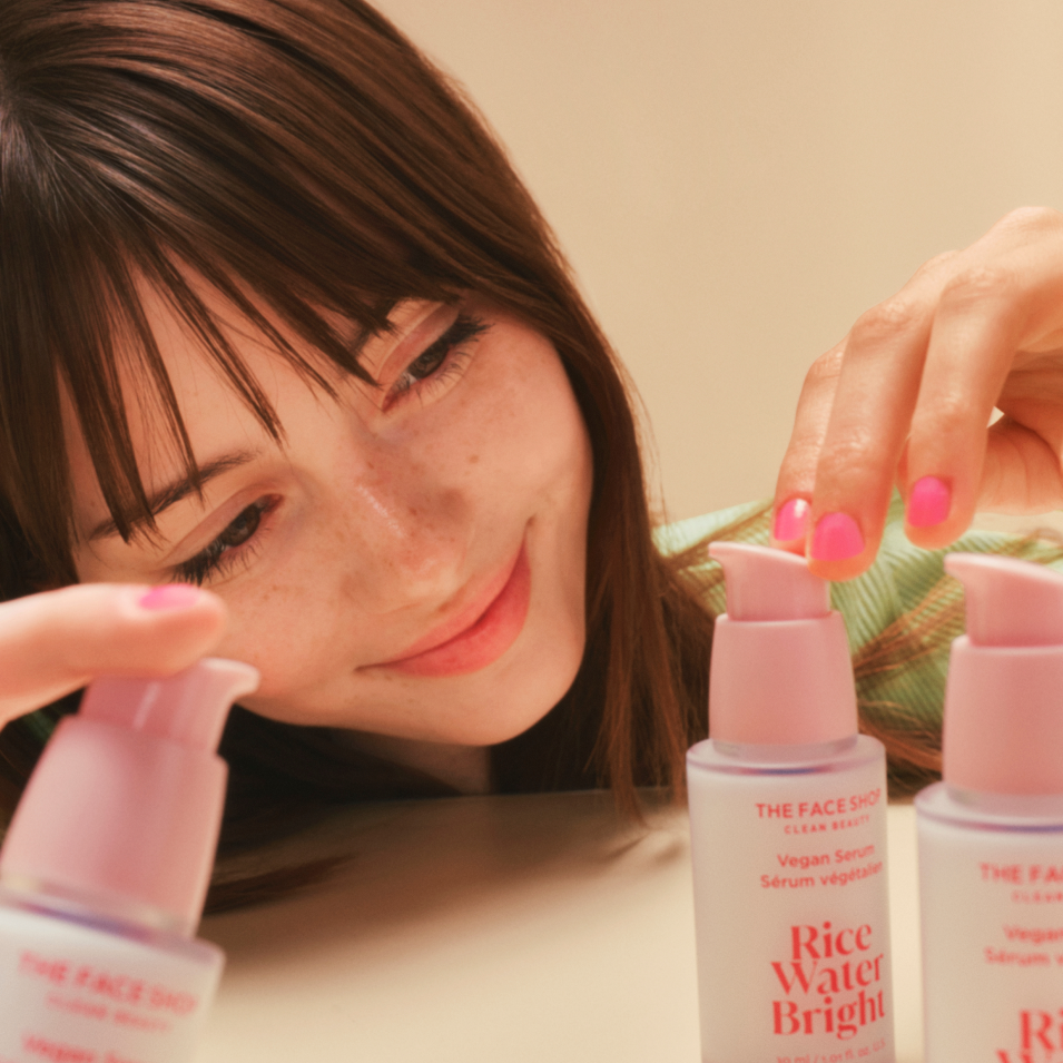 A woman with bangs smiles gently as she examines The Face Shop Vegan Serum on a table.