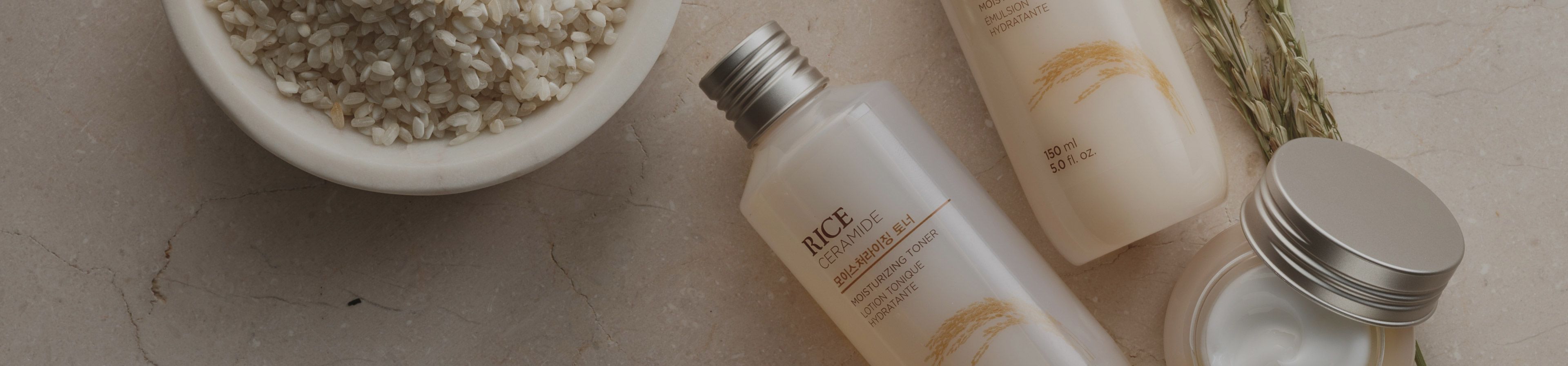 Two bottles and a jar of rice ceramide skincare products on a textured surface with rice grains and stalks nearby, emphasizing natural ingredients.