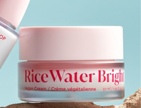 A pink-lidded jar of "Rice Water Bright Vegan Cream" sits on a brown surface against a light blue background.