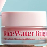 A pink-lidded jar of "Rice Water Bright Vegan Cream" sits on a brown surface against a light blue background.