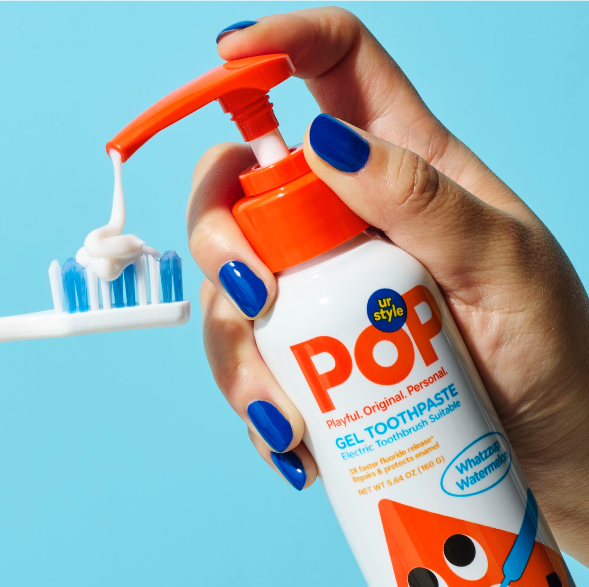 A hand holding a white bottle of POP Kids Gel Whatzzup Watermelon Toothpaste, pumping the toothpaste out on a white-and-blue toothbrush ona a light blue background.