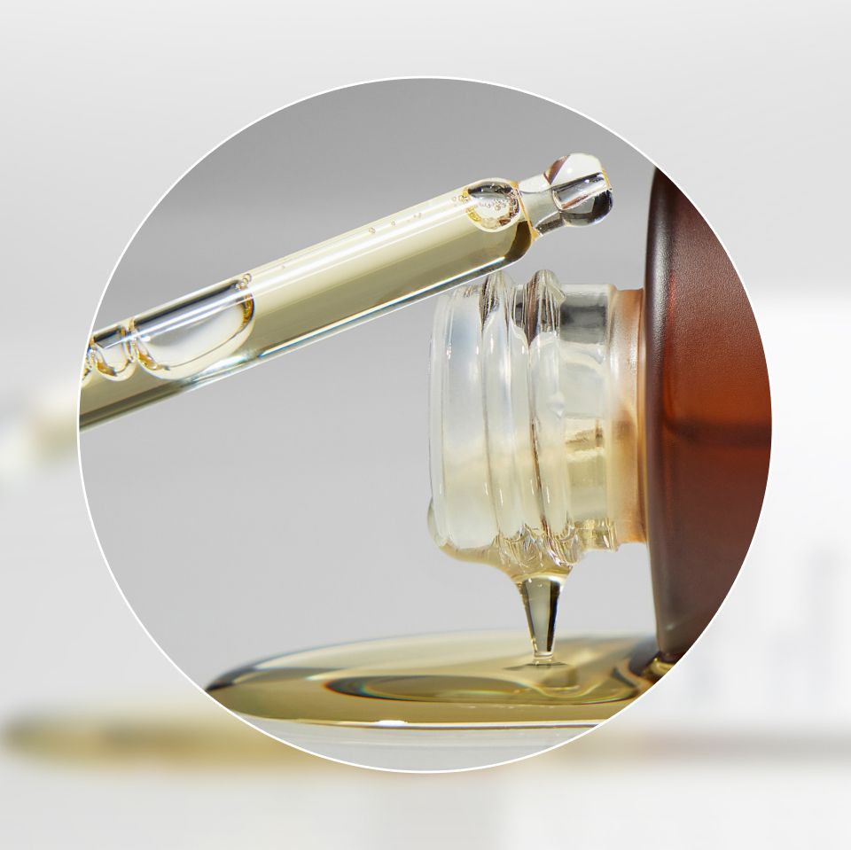 Close-up image of a glass dropper dispersing oil from a brown glass bottle. The dropper is releasing a drop of golden liquid into the bottle, with a smooth and shiny texture evident. The background is blurred, focusing attention on the dropper and bottle.