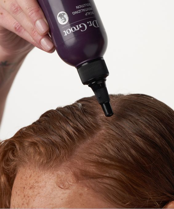 A hand applies Dr. Groot Scalp Revitalizing Solution from a purple bottle to a person's reddish-brown hair and scalp.