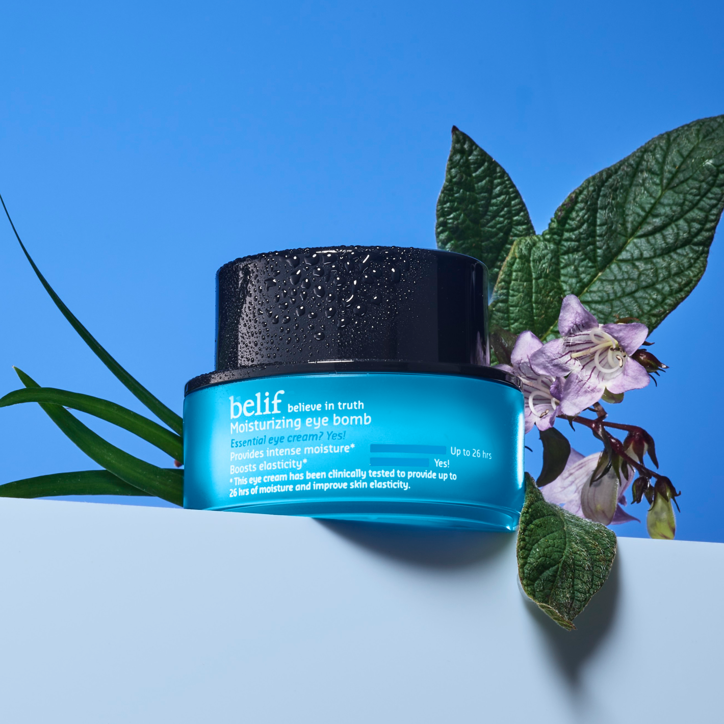 A bottle of the belif Moisturizing eye bomb with purple flowers on a blue background.