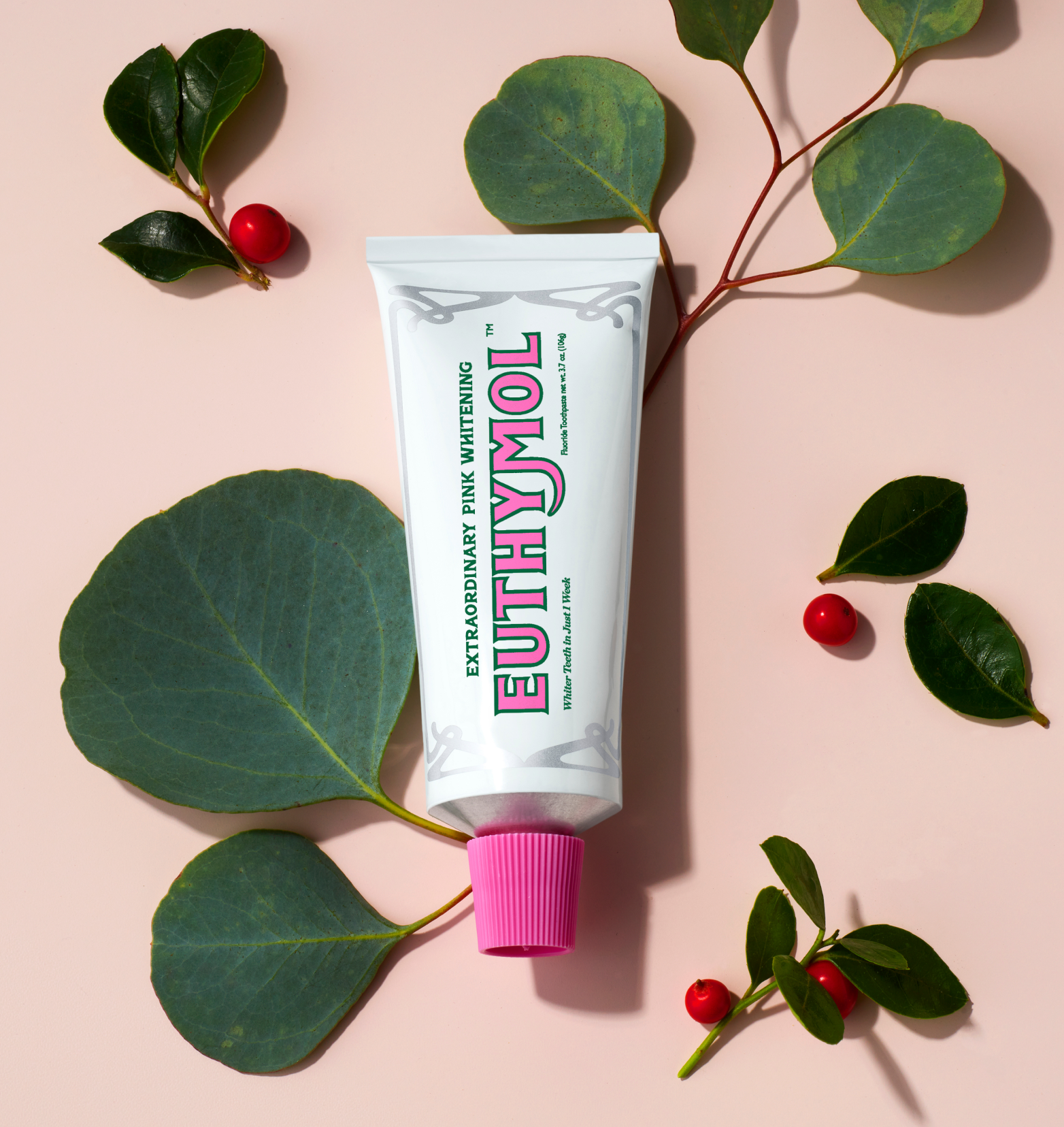 A white tube of Euthymol Whitening Toothpaste, lying on a light pink surface with leaves and cranberries around.
