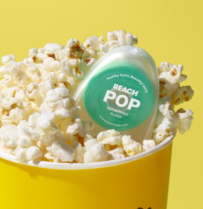 A container of Reach POP Peppermint Floss sits atop a bucket of fluffy popcorn.