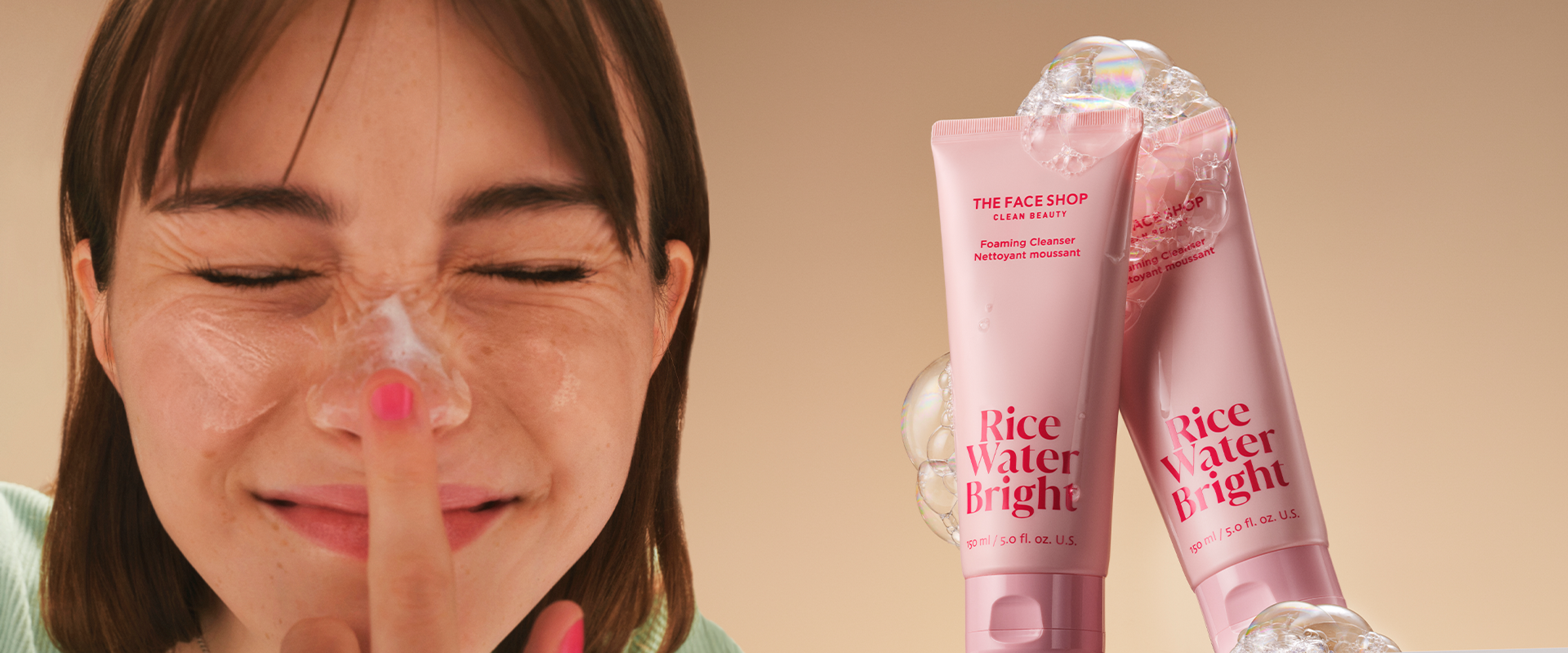 A woman with closed eyes touches her nose, smiling gently. in the background, two tubes of the face shop rice water bright cleansing foam covered in bubbles are displayed with a light glow.