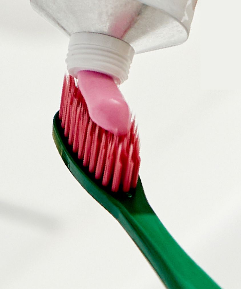 A close-up of a green Euthymol Original Regular Toothbrush with dark pink bristles, as Euthymol Toothpaste is being squeezed onto it from a tube.