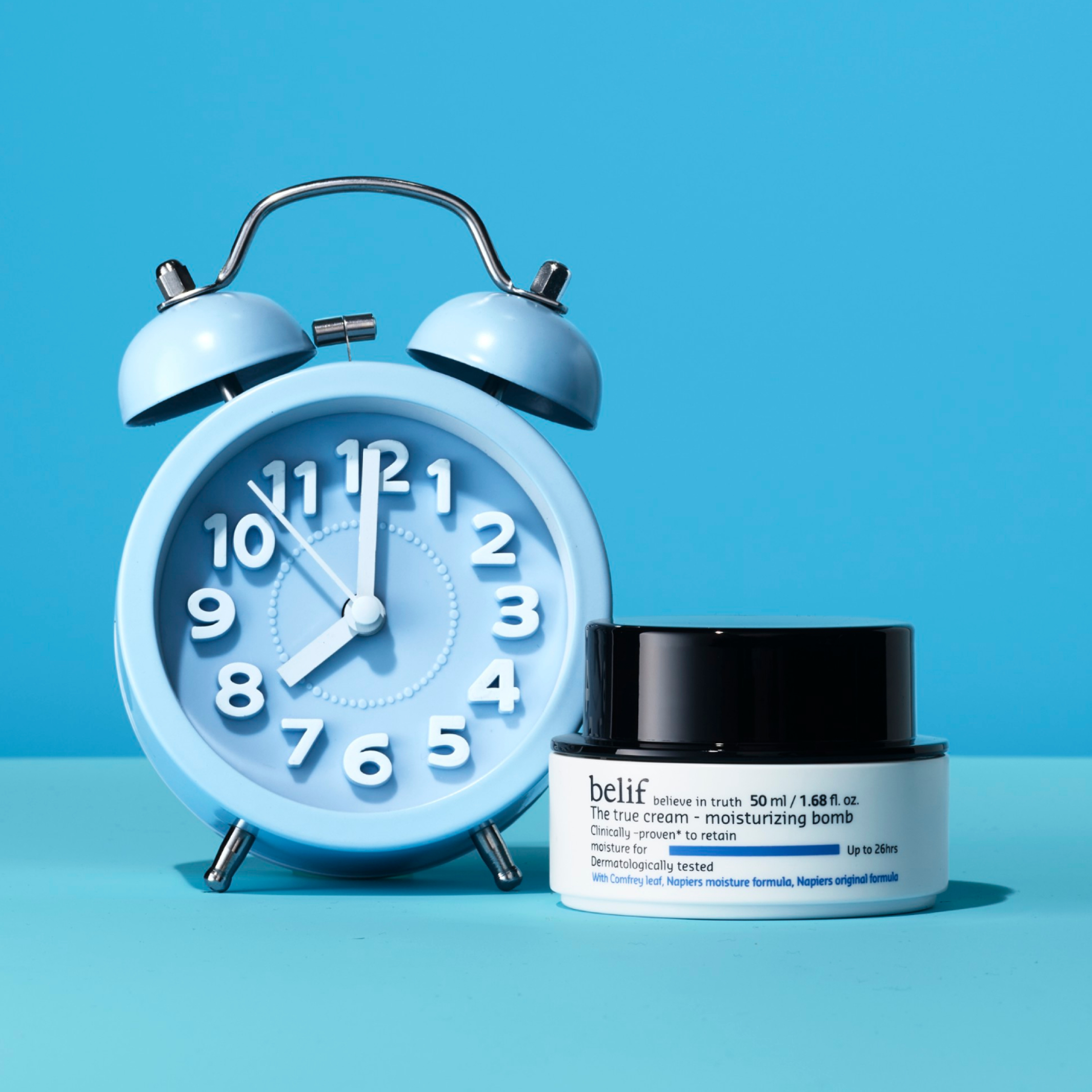 A jar of belif The True Cream Moisturizing Bomb standing on a light blue surface with a blue analog alarm clock standing by its side.