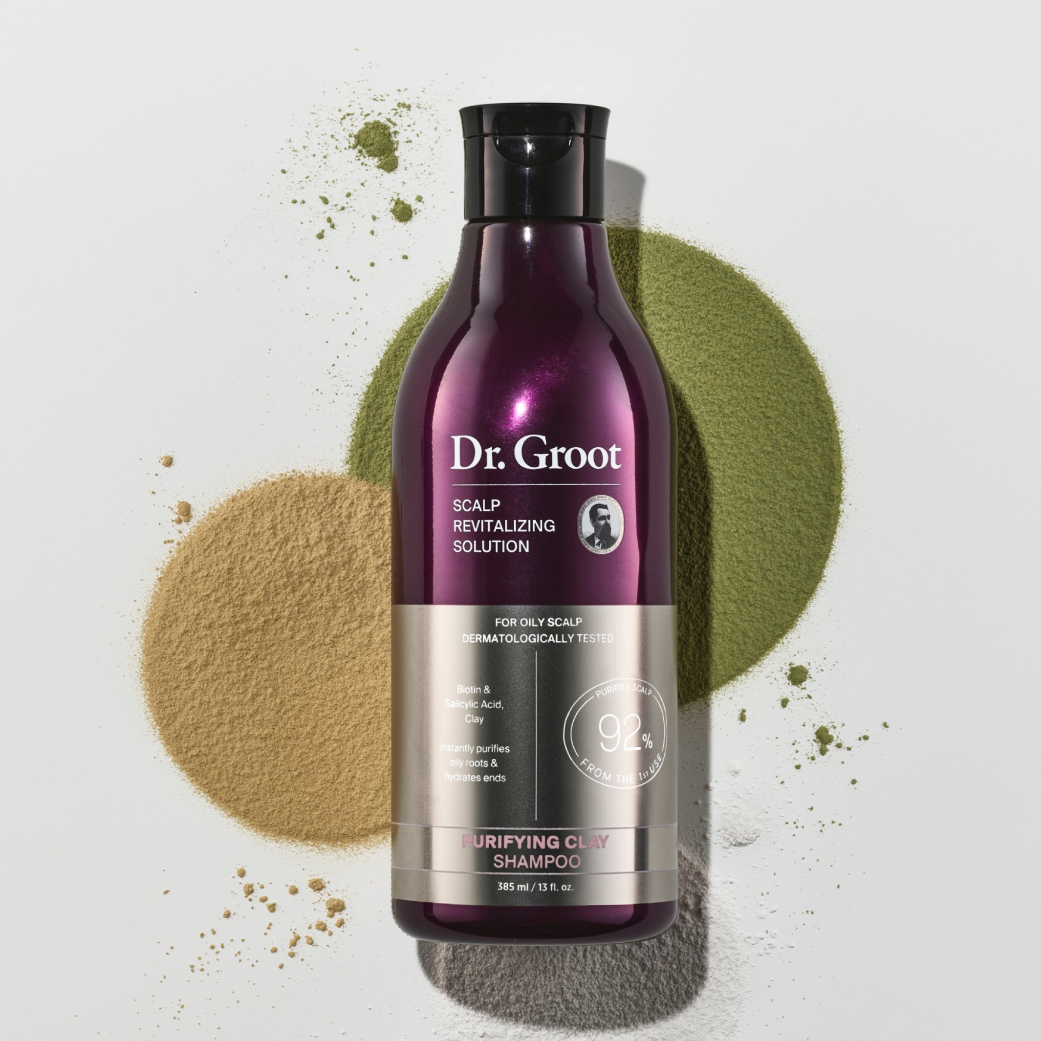 A dark purple bottle of Dr. Groot Purifying Clay Shampoo lying on a grey surface in three beige, green and white circle of clay powder.