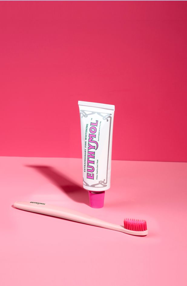 A portrait of a Euthymol Whitening pink toothbrush in front of  a tube of Euthymol Whitening Toothpaste on a light pink surface with a dark pink background.