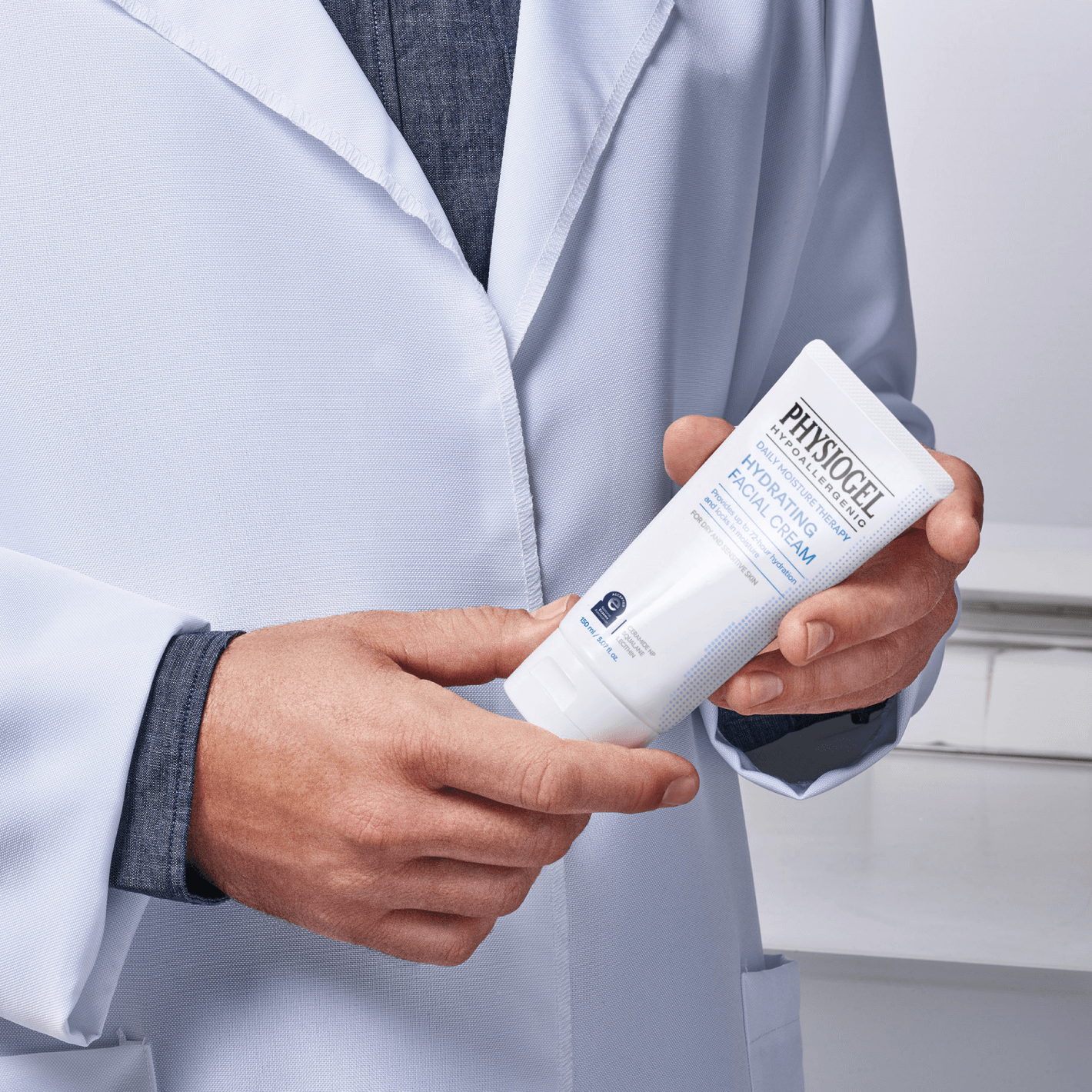 A person in a lab coat holds a tube of Physiogel Hypoallergenic Daily Moisture Therapy facial cream.