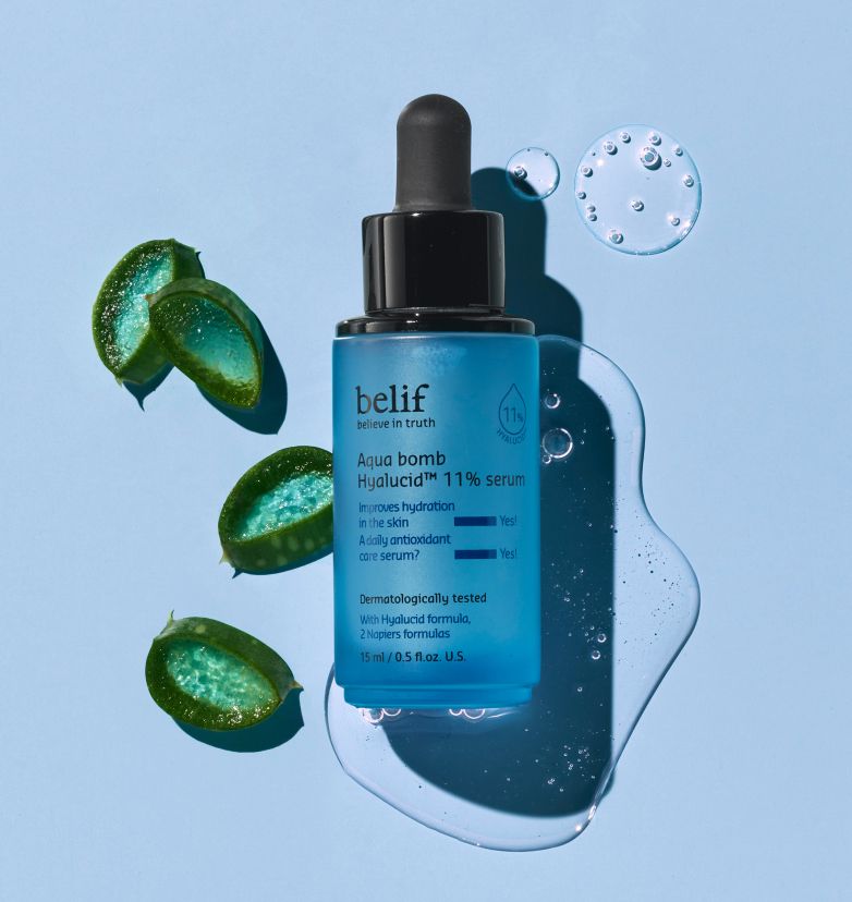 A tiny blue bottle of belif Aqua Bomb Hyalucid 11% Serum lying in a tiny puddle of the serum with pieces of aloe scattered around