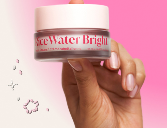 Hand holding a "rice Water Bright Vegan Cream" jar against a pink and white background with floating pink and white rice grains.