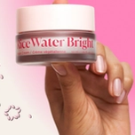 Hand holding a "rice Water Bright Vegan Cream" jar against a pink and white background with floating pink and white rice grains.