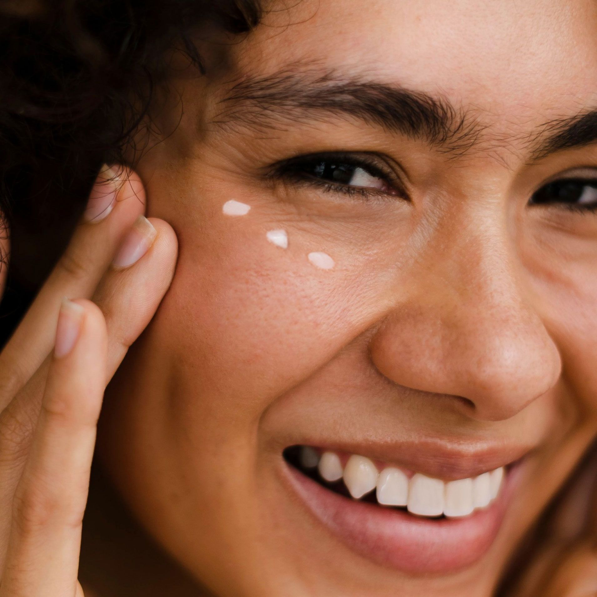 Close up of a model smiling and touching her face with three dots of eye cream under her eye.
