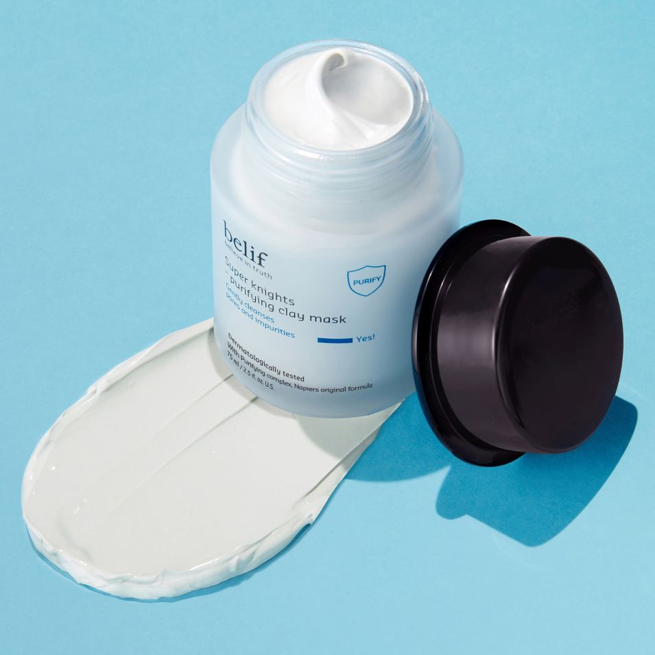 A white jar of belif Super Knights Purifying Clay Mask standing on a light blue surface on a spread of the mask