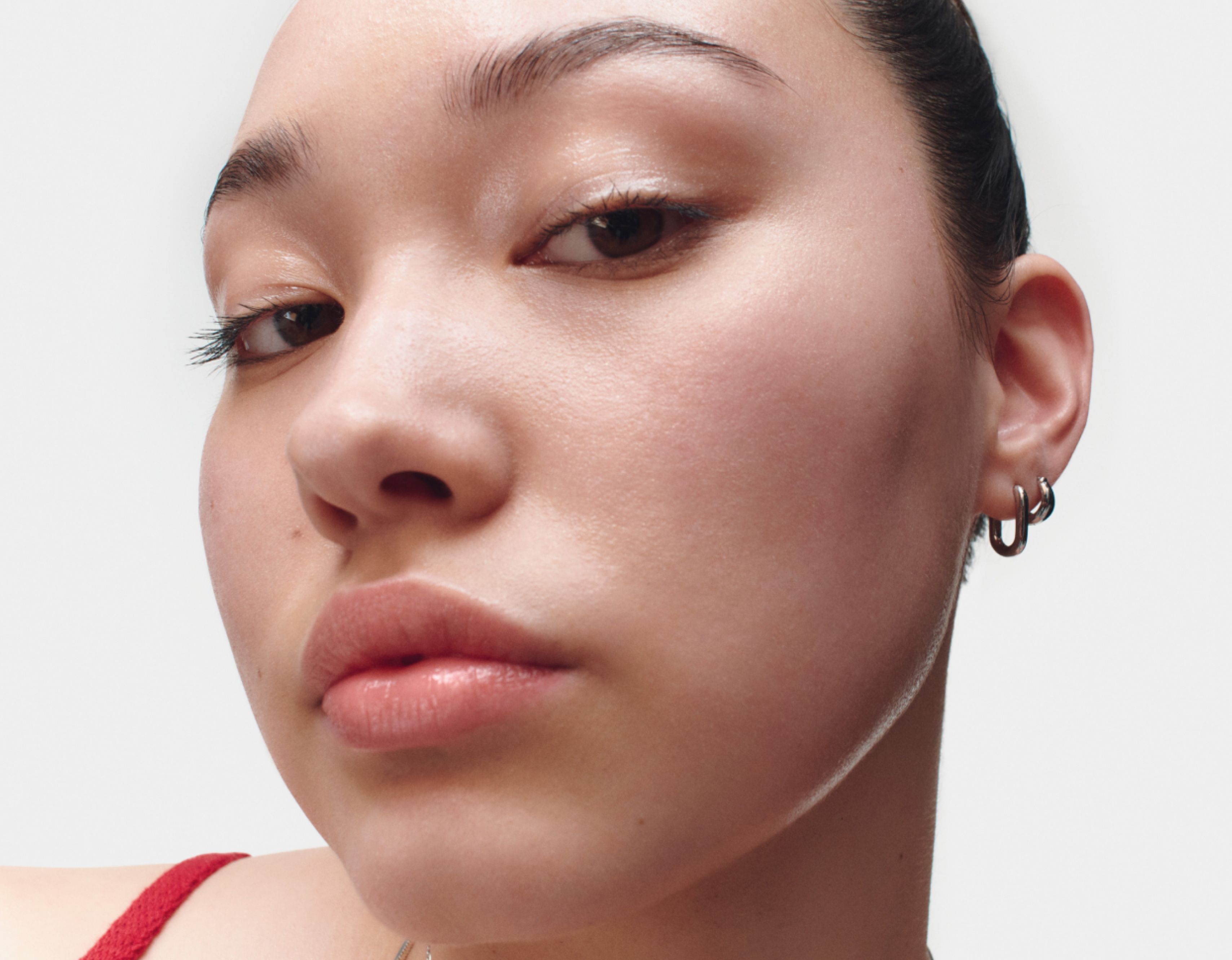 Close-up shot of a person with smooth skin, wearing subtle makeup. They have dark, neatly groomed eyebrows and are looking directly into the camera with a neutral expression. The person is wearing small hoop earrings and a visible red strap from their clothing. The background is plain white.