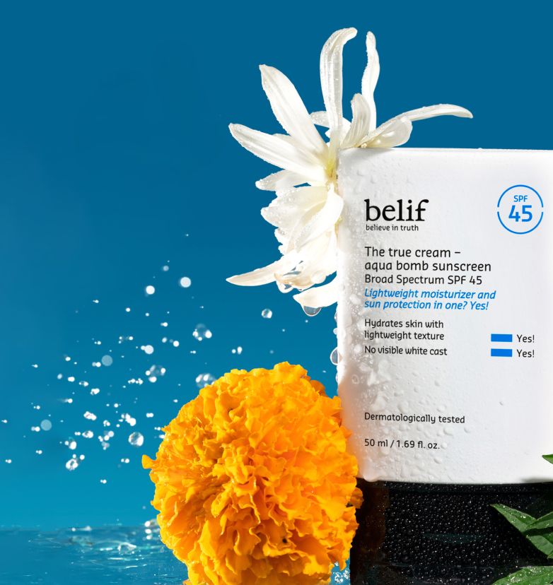 A tube of belif Aqua Bomb SPF 45 Sunscreen drenched in water droplets, surrounded by misty flowers on a blue background.