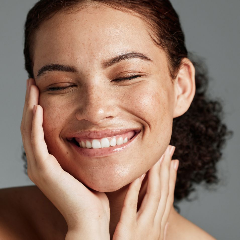 A model smiling with her eyes closed touching her face and neck with her hands
