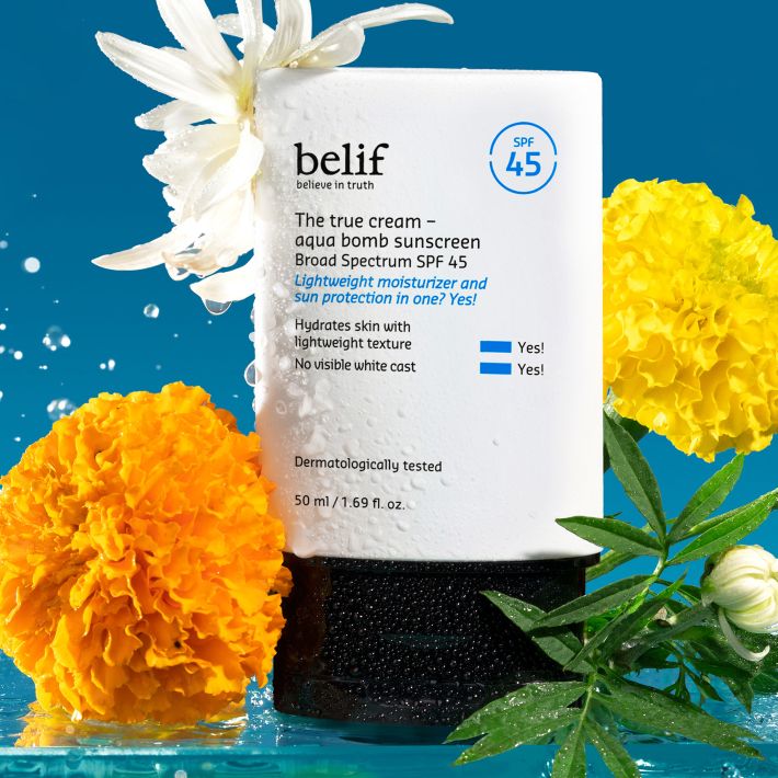 A tube of belif Aqua Bomb SPF 45 Sunscreen drenched in water droplets, surrounded by misty flowers on a blue background.