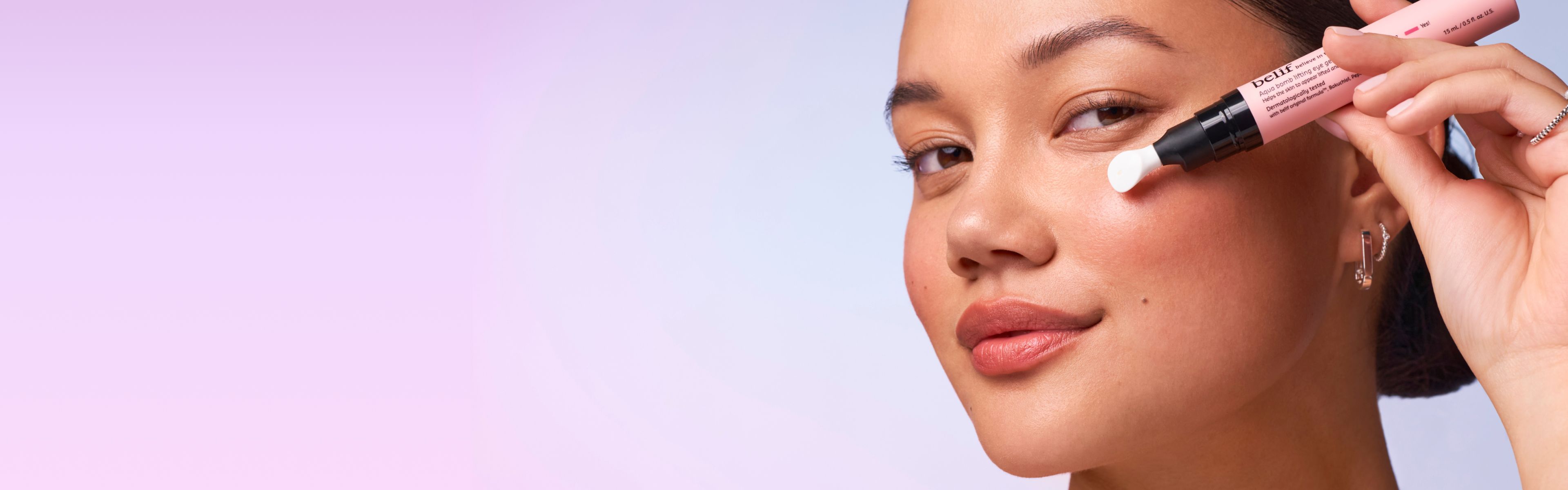 A woman applies belif Aqua Bomb Lifting Eye Gel with a white applicator from a pink tube to her undereye.