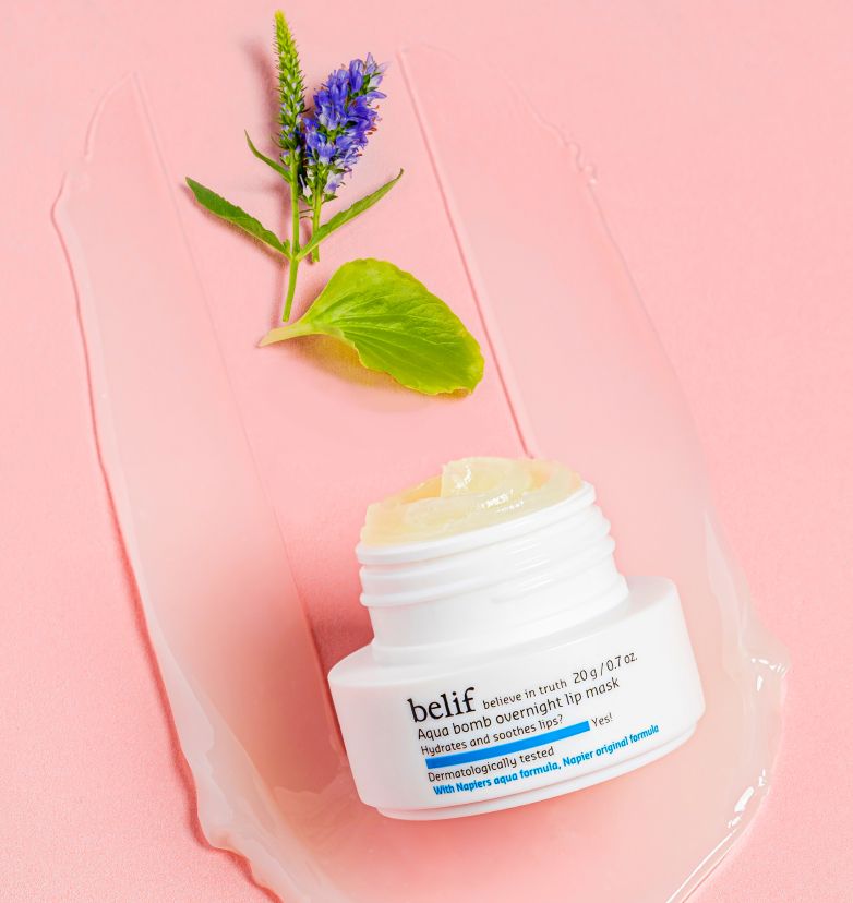 A white jar of belif Aqua Bomb Overnight Lip Mask lying in a spread of the mask on a light pink surface with a blue flower lying around